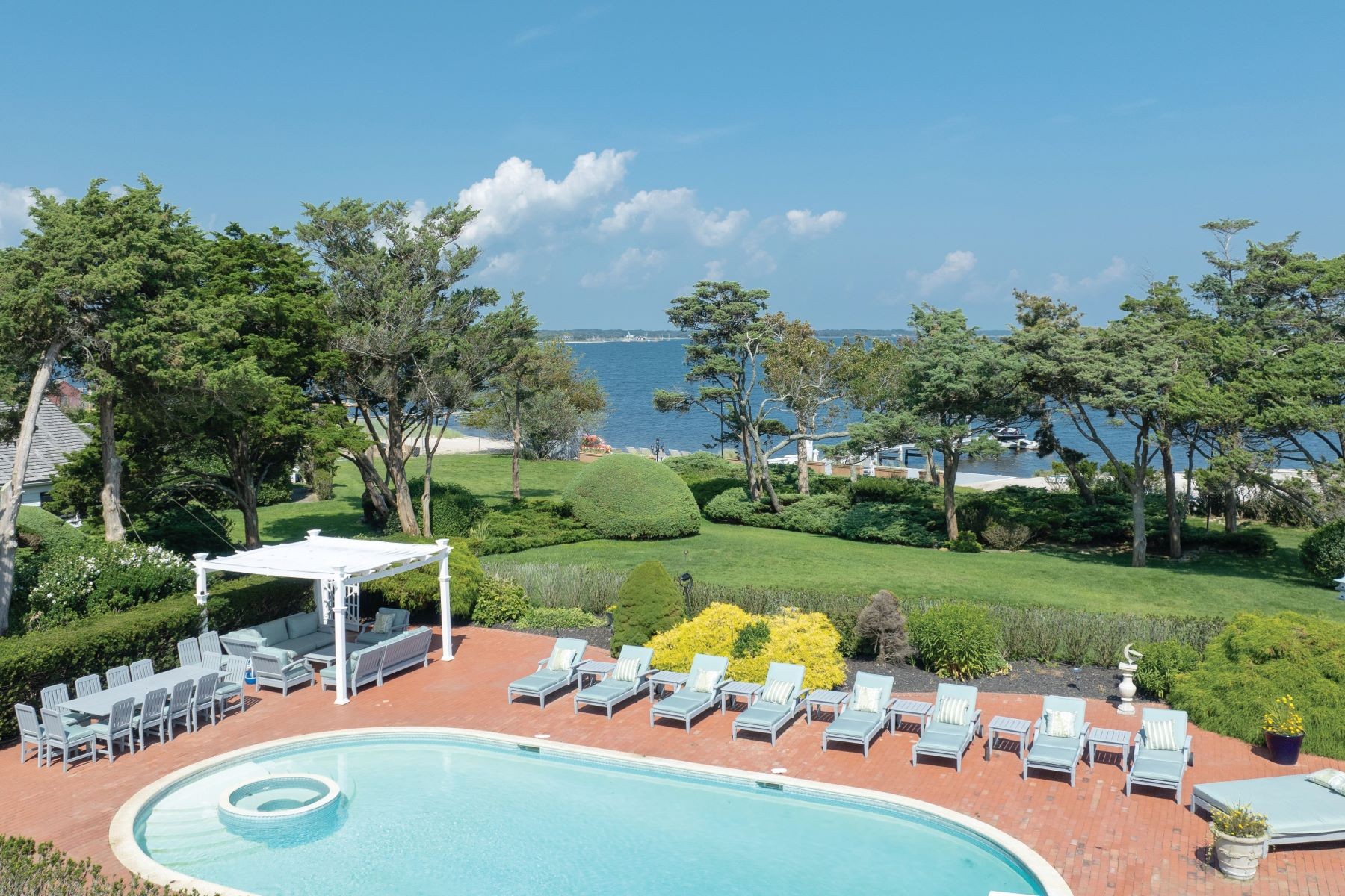 The waterside pool terrace at 16 Point Road, a white pergola and rows of cushioned chaises on brick, framed by lawn and trees with the bay beyond