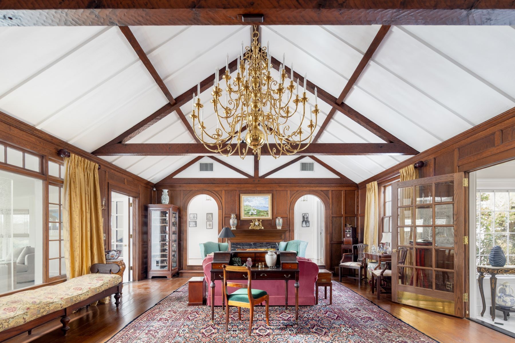 The vaulted great room at 16 Point Road with original exposed beams, mahogany-paneled walls, twin arched alcoves flanking the fireplace, and a monumental gilt-brass chandelier