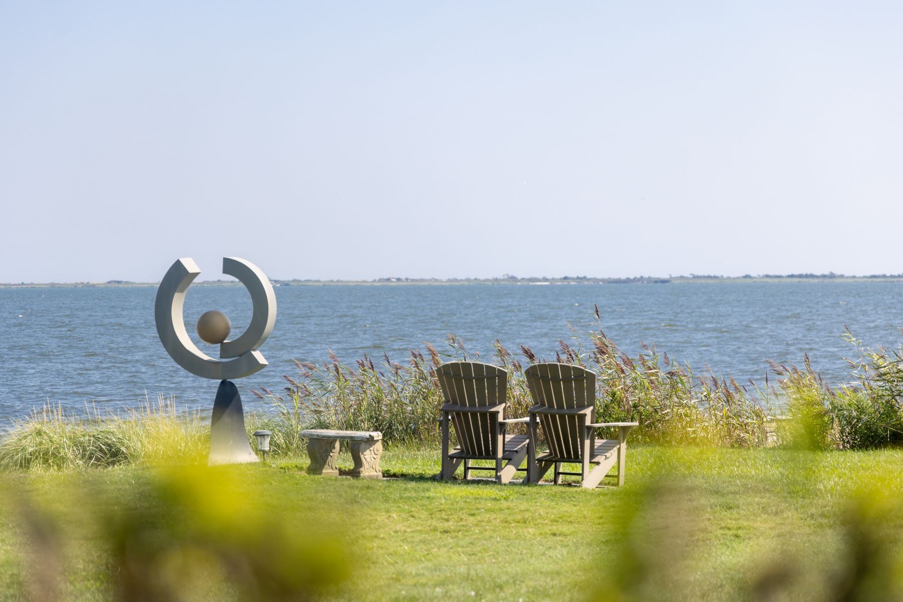 A modern bronze sculpture on the lawn at 16 Point Road beside two wooden Adirondack chairs and a stone bench, facing the open water of Great South Bay