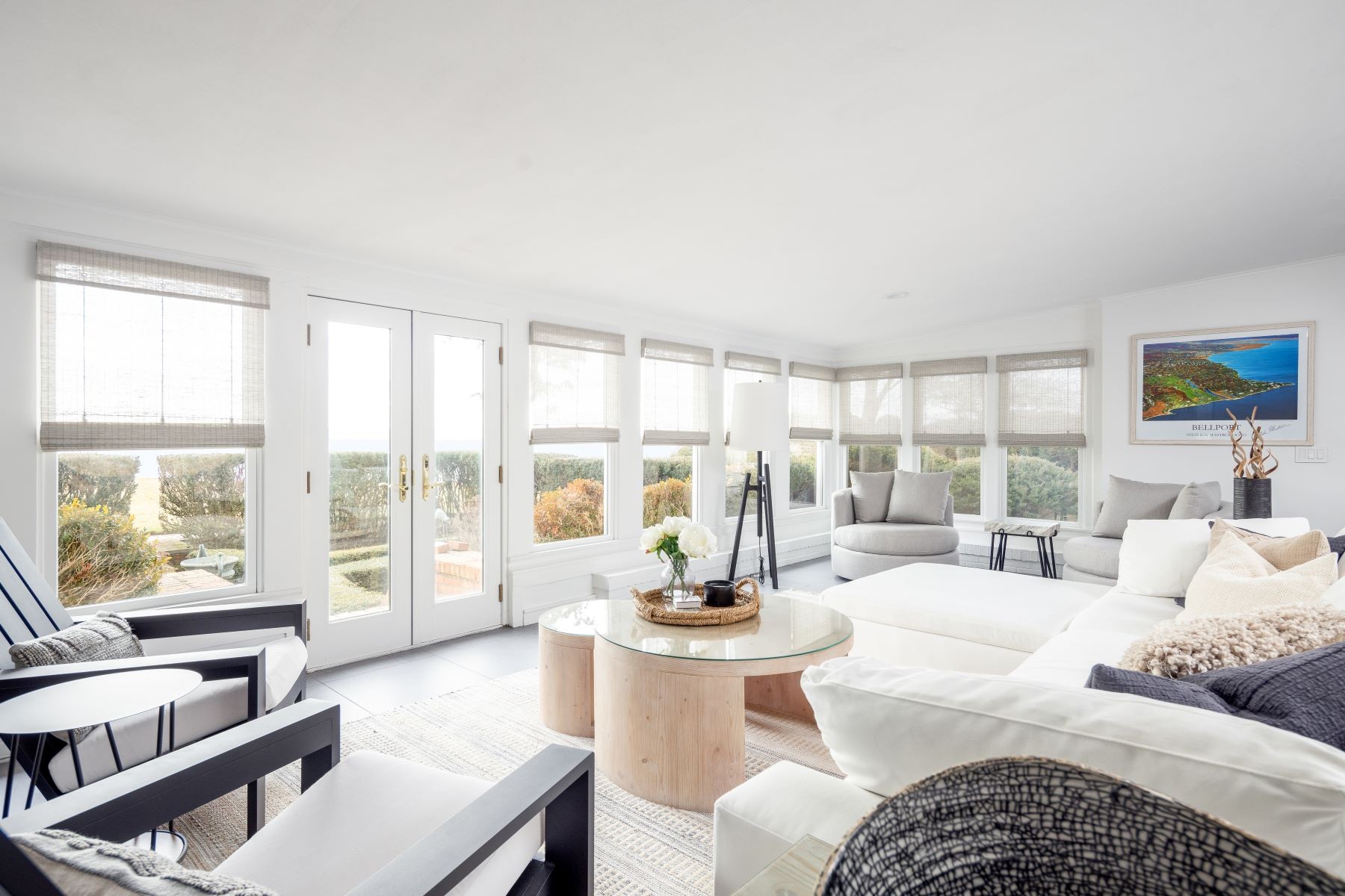 A sunlit living room at 16 Point Road in soft whites, walls of windows and French doors opening to the lawn and water, a round wood coffee table at the center