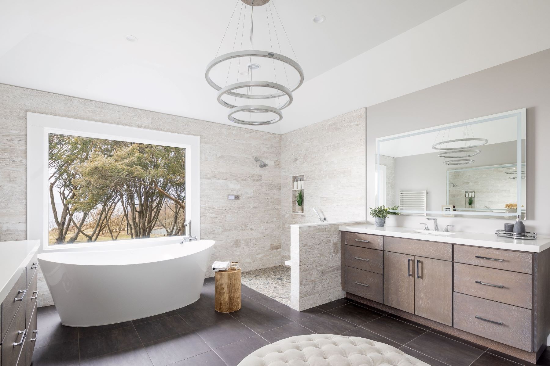 The primary bath at 16 Point Road with a freestanding soaking tub under a picture window, a triple-ring chandelier, a walk-in stone shower, and a floating oak vanity over dark tile