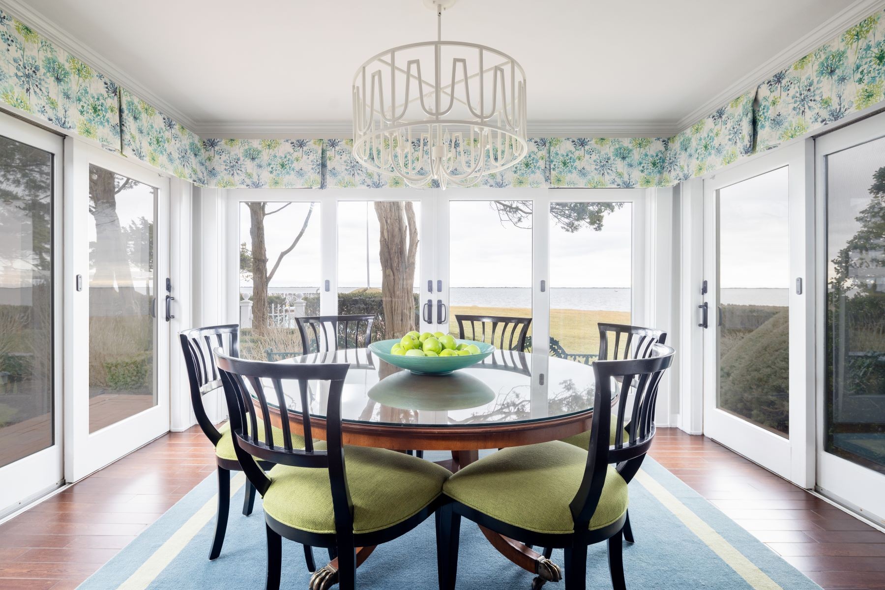 A breakfast room at 16 Point Road wrapped on three sides by glass doors that open to the lawn and the bay, a round pedestal table with green-cushioned chairs at the center