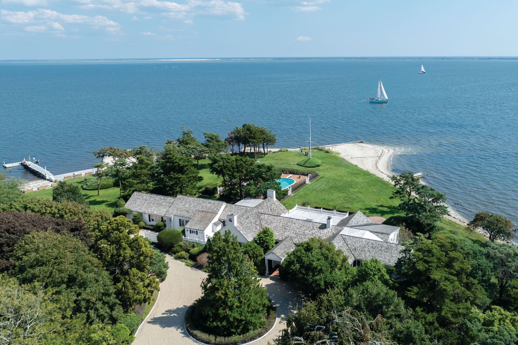 Aerial of 16 Point Road, a white shingle-style estate alone on a private wooded point with Great South Bay wrapping it on three sides, a dock at left and a sandy beach at right